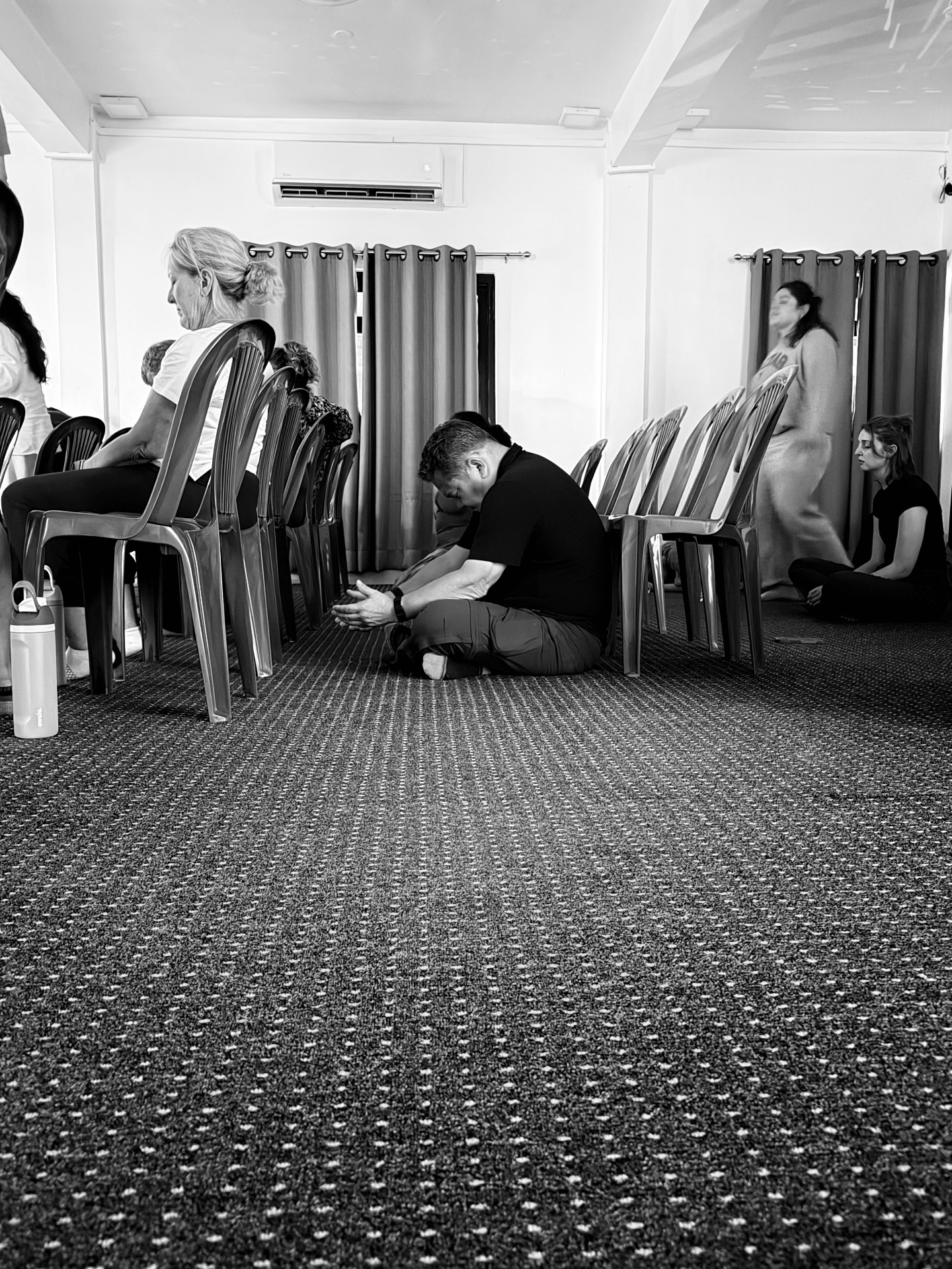 A Racer kneeling on the floor in prayer beside rows of chairs during a worship gathering — black and white