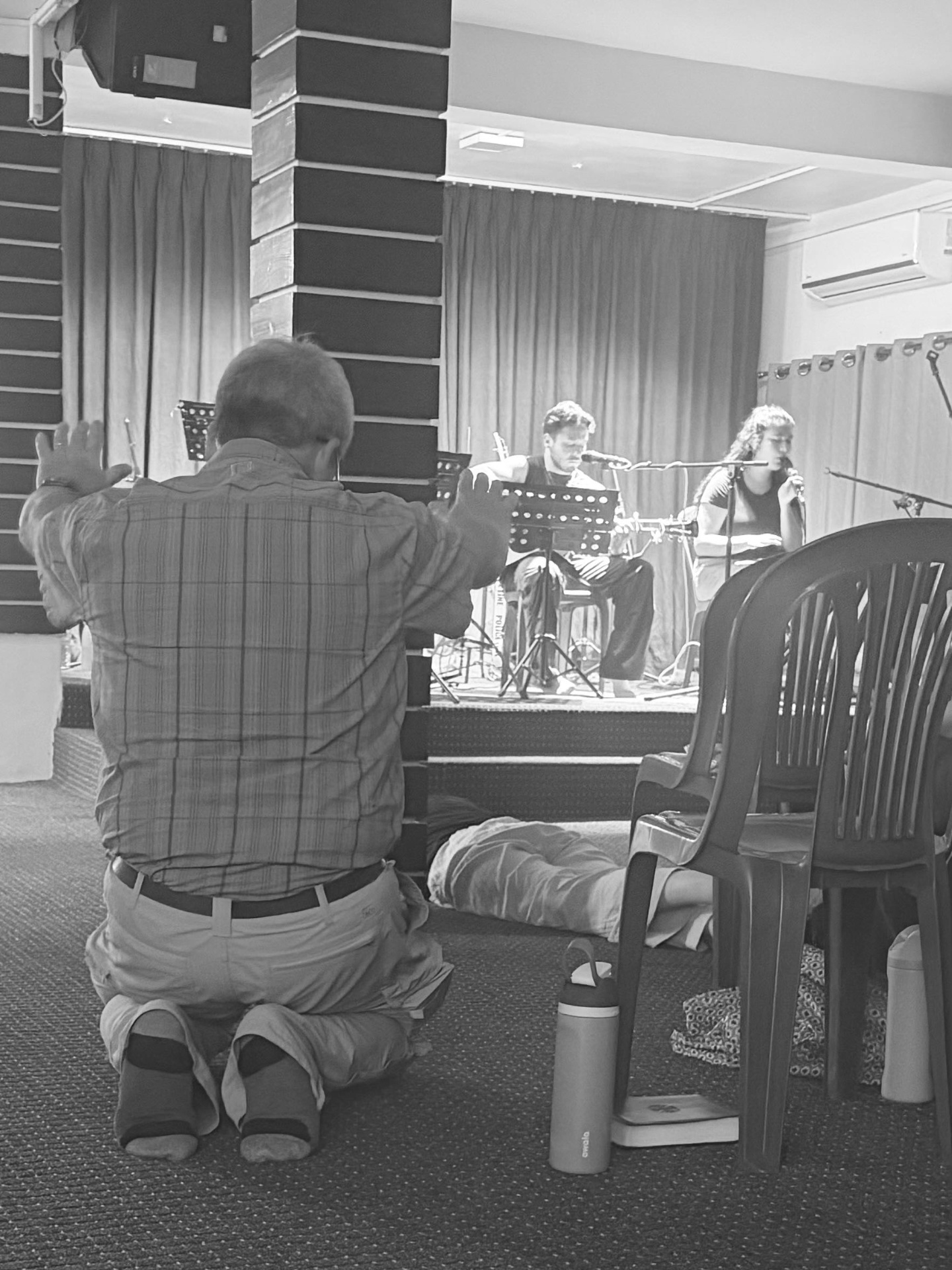 A parent kneeling in worship with hands raised while a worship band leads from the stage — black and white